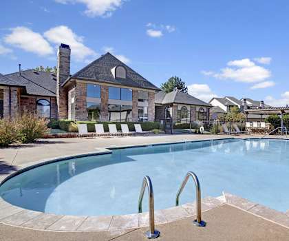 The large, resort style, light blue outdoor pool with white pool chairs, tables, and chairs with the apartments in the background at Stoneleigh on May Apartment Homes. 