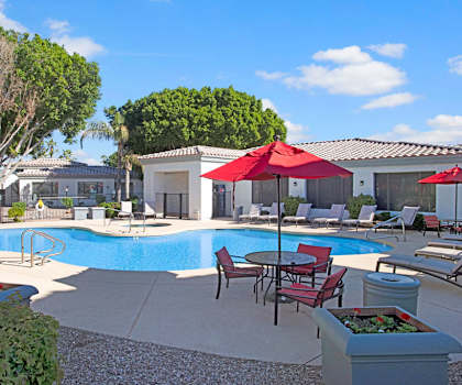 The turquoise outdoor pool with pool chairs, tables with red umbrellas over them, and chairs with the apartments and main office building in the background at Pala Mesa Apartments.