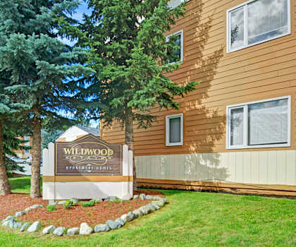 The exterior sign with trees and surrounded by greenery with the buildings in the background of Wildwood Estates Apartment Homes. 