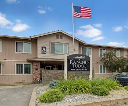 The exterior sign with trees and surrounded by greenery with the buildings and an American Flag in the background of Rancho Tudor Apartments. 