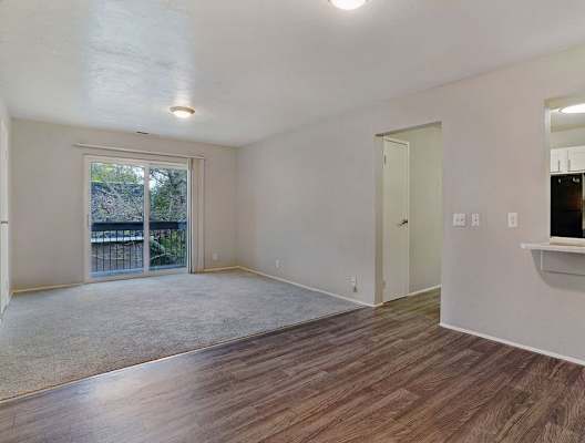 A room with a sliding glass door leading to a balcony, a window, and a kitchenette with white cabinets.