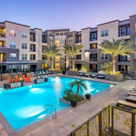 A large, turquoise, resort-style swimming pool surrounded by palm trees in front of an apartment building at the Flats at San Tan.