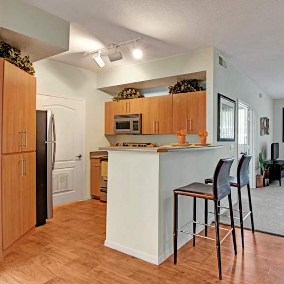 The interior of an apartment with showing the kitchen with a refrigerator, microwave, oven, barstool chairs, a coffee table, a couch, chairs, and a TV at Acclaim Apartments. 