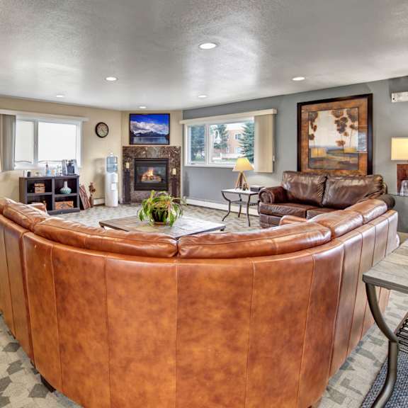 A lounge with a U-shaped brown leather couch, a darker brown leather couch, wall art, two windows, shelves, a coffee table, and a fireplace at the Club at Eagle Point Apartments.