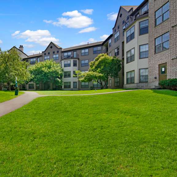 The stunning pathways surrounded by grass and trees leading to the gray apartment building at Fairways Apartment Homes. 