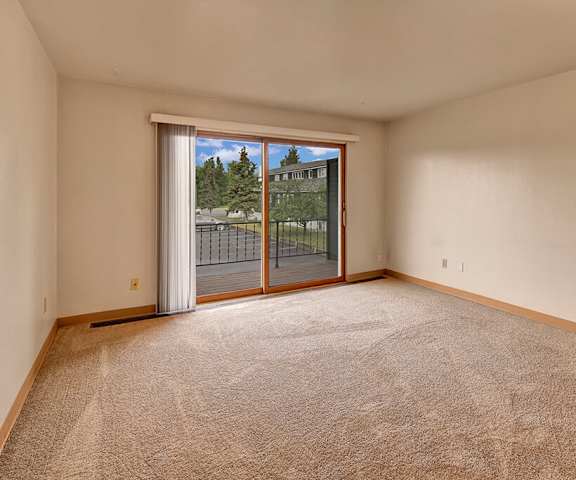 A large, empty living room with carpeting and a sliding glass door to the balcony at the Alaskan Apartment Homes. 