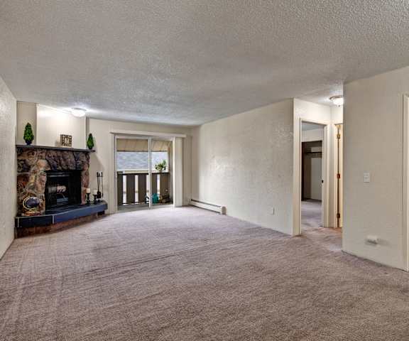 A large, empty living room with carpeting, a fireplace, overhead lighting, and a sliding glass door to the balcony at the Greenbriar Apartment Homes. 