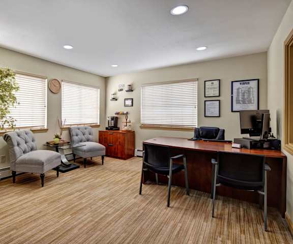 The leasing office with a desk, a desktop computer, chairs, gray plush chairs, a coffee station, a plant, three windows, and wall art at the Greenbriar Apartment Homes. 