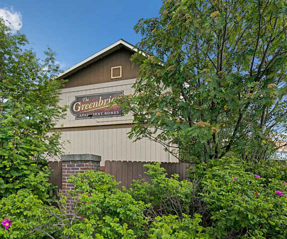 The exterior brown sign on the side of a building with trees and surrounded by greenery, trees, and flowers with the buildings in the background of the Greenbriar Apartment Homes. 