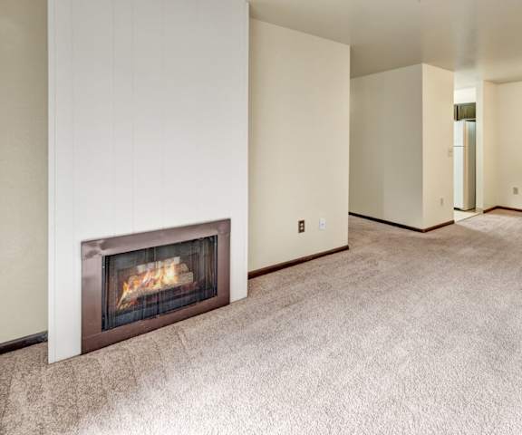 An empty living room with carpeting, fireplace, and a sneak peak of the kitchen at Ladera Villa Apartments. 