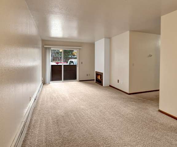 A view of the empty carpeted living room with a window and the kitchen with a door to the balcony at Ladera Villa Apartments. 