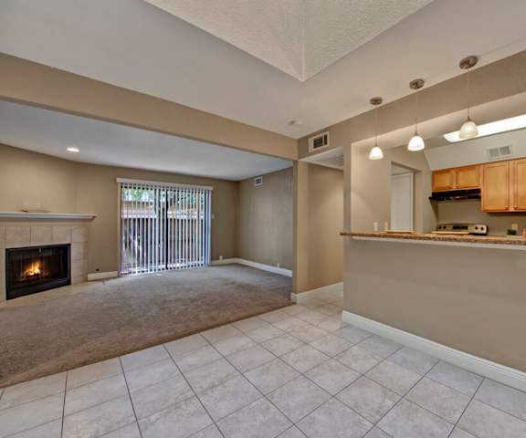 A view of the empty carpeted living room with a door to the patio, fireplace, and the kitchen with white appliances and wooden large cabinets at Las Colinas Apartments. 