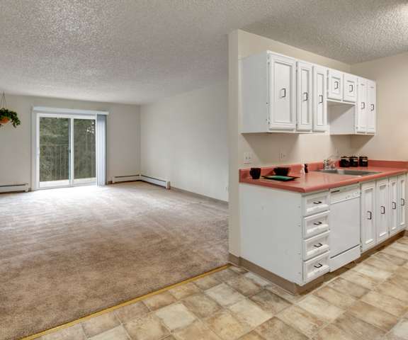 A view of the empty carpeted living room with a glass door to the balcony, and the kitchen with white appliances and wooden large cabinets at Quiet Creek Apartment Homes. 