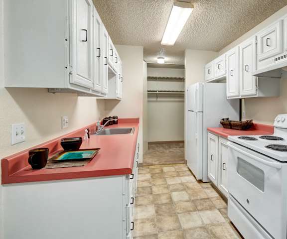 A cozy kitchen with white appliances, a red counter with lots of room, and spacious white wooden cabinets at Quiet Creek Apartments. 