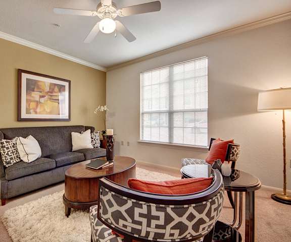 The spacious living room with a dark gray couch, white throw pillows, a large window, overhead lighting with a ceiling fan, wall art, two orange and brown plush chairs, coffee table, a lamp, and decorations at the Residence at Heritage Park Apartment Homes. 