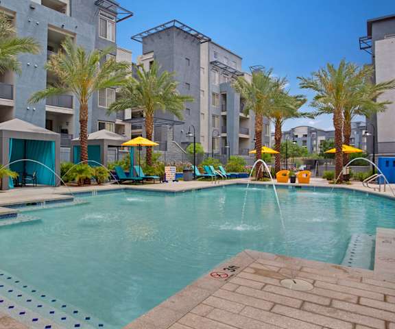 The large, turquoise outdoor pool surrounded by palm trees, pool chairs, tables, cabanas, and chairs with the apartments in the background at Rio Paradiso Apartment Homes. 