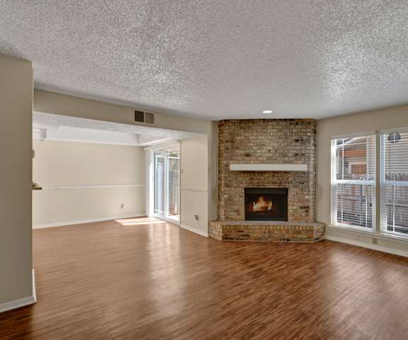 An empty living room with hardwood floors, windows, a fireplace, and a sliding glass door to the patio at Summertree Place Residences. 