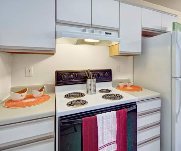 A cozy kitchen with white appliances, kitchen utensils, overhead lighting, light countertops with lots of room, a bar counter with barstools, and large wooden cabinets at Timber Ridge Apartment Homes. 