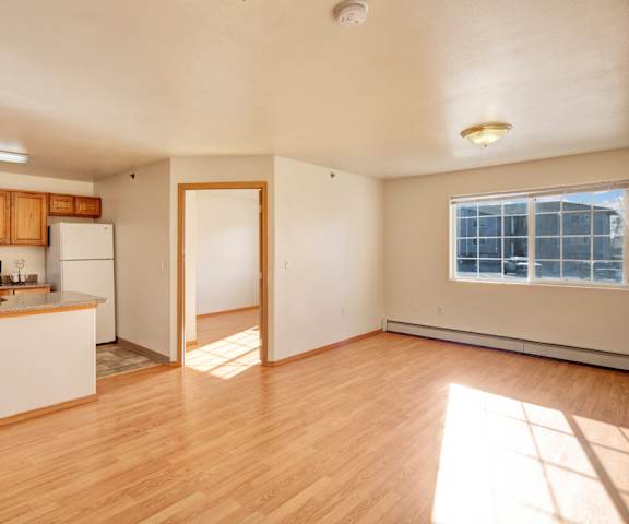 A large, empty living room with hardwood flooring and kitchen with a door to a balcony at Timber Ridge Apartment Homes. 