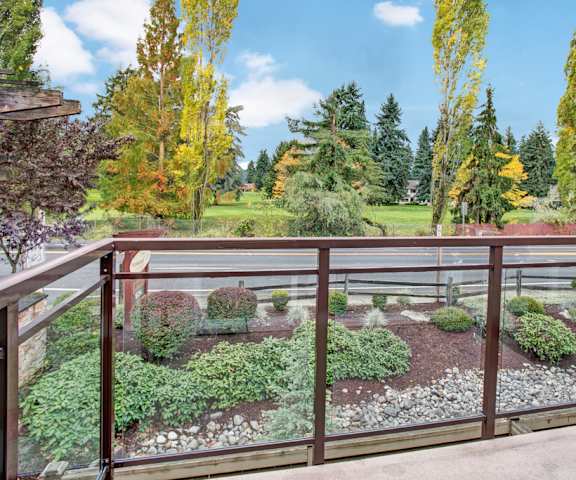 A balcony with glass panels overlooking the garden below, and across the street, a large field of grass surrounded by trees at Vermont Apartment Homes. 