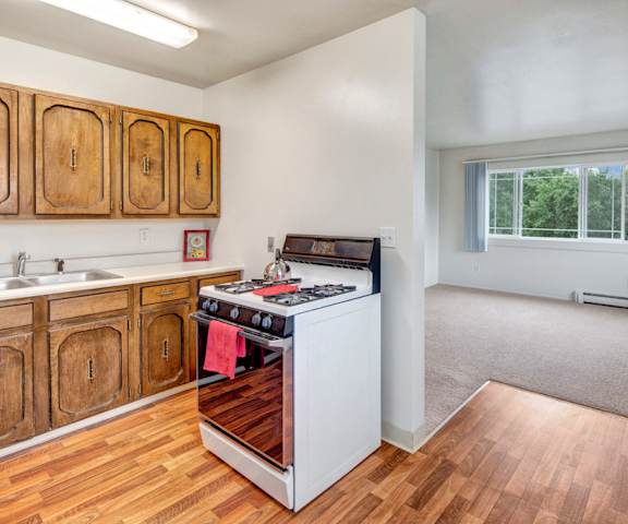 A spacious/cozy kitchen with white appliances, kitchen utensils, overhead lighting, light countertops with lots of room, a bar counter with barstools, and large wooden cabinets at the Village at Calais Apartment Homes. 
