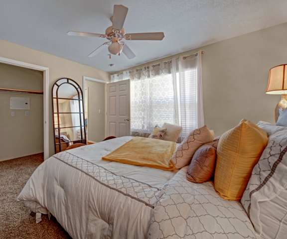 A spacious bedroom with a queen-size bed with a white and yellow bedspread, a dresser, wall art, lamps, decorations, a window with white lace curtains, a closet, and nightstands with lamps at Summit Pointe Apartment Homes. 