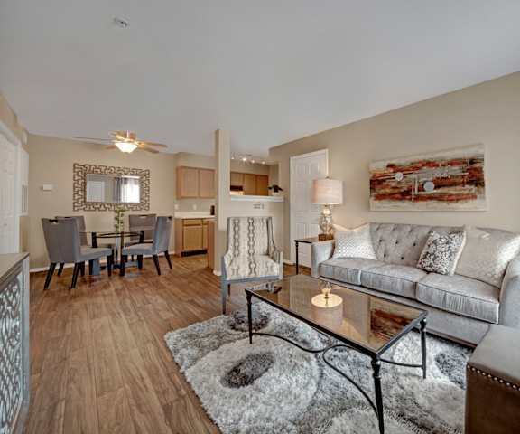 The spacious living room with a gray couch, a plush chair, glass coffee table, overhead lighting with a ceiling fan, decorations, a TV, and view of the dining room with a table, gray chairs, and place settings for four, and the kitchen at Summit Point Apartment Homes. 