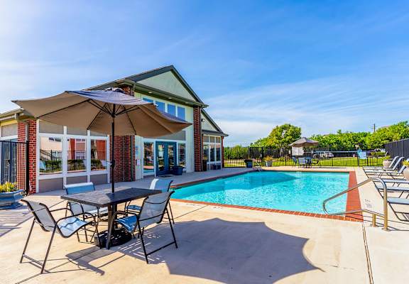 A poolside table and chairs are set up under an umbrella next to a swimming pool.