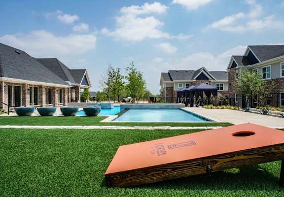 A giant game of cornhole is in the foreground of a residential area with houses and pools.