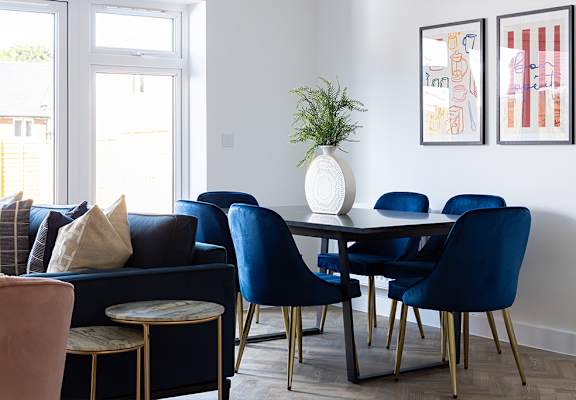 A living room with a dark blue sofa, a round pink chair, a white vase with a green plant, and two framed artworks on the wall.