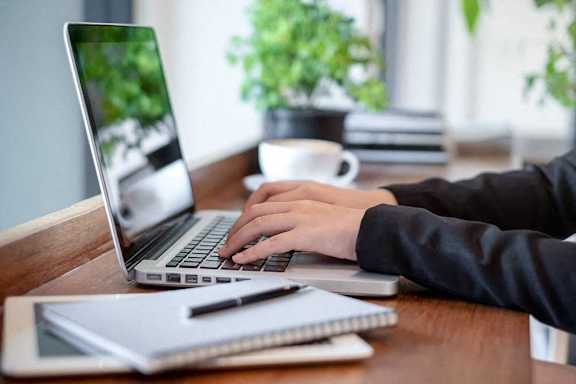 A person is typing on a laptop with a notebook and pen beside them.