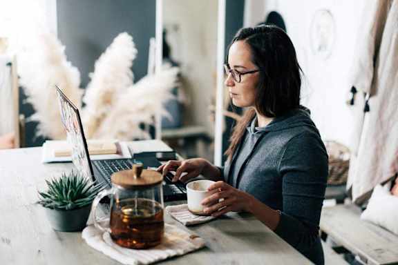 A woman is sitting at a table with a laptop and a cup of tea.