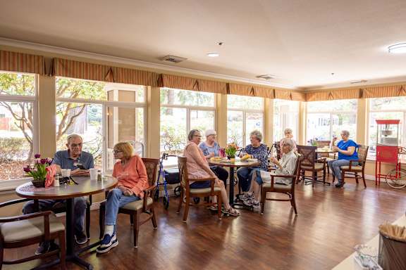 A group of people are sitting around tables in a room with wooden floors and large windows.