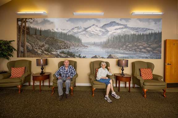 Two elderly people sitting in chairs in front of a mural of a mountain landscape.