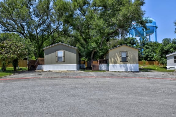 a parking lot in front of two sheds with trees and a water tower in the background at Buckingham Village in Universal City, TX