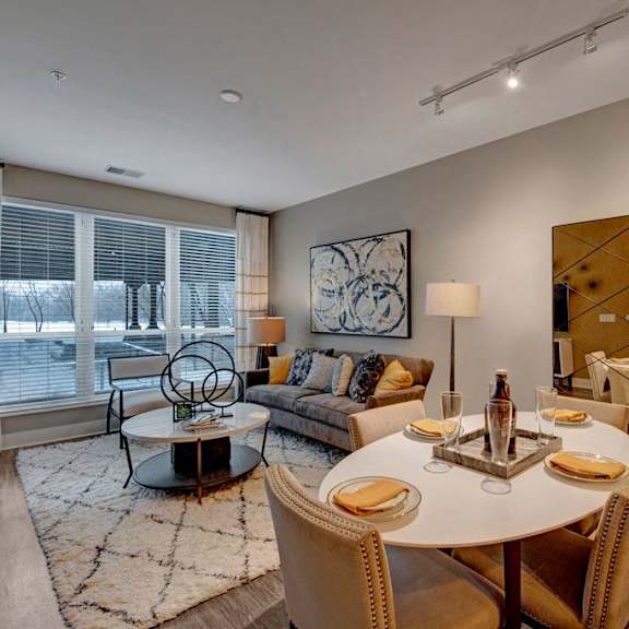 The spacious living room with a plush couch with colorful throw pillows, plush chairs, large windows with white curtains, coffee table, overhead lighting with a ceiling fan, decorations, a TV, and view of the dining room with a table, chairs, and place settings for four at River House Apartment Homes.