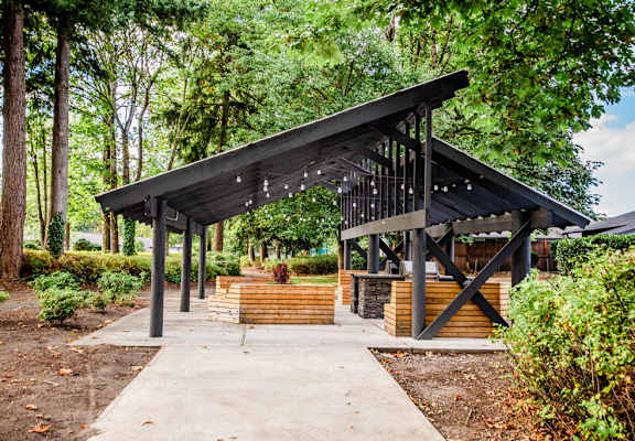 A pavilion with a black roof and wooden pillars is surrounded by trees and bushes.