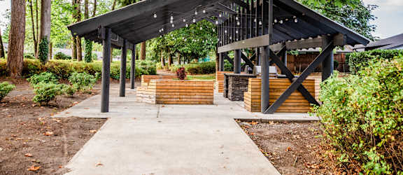 A pavilion with a black roof and wooden pillars is surrounded by trees and bushes.