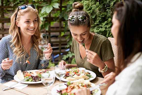 three women sitting at a table eating food and drinking wine