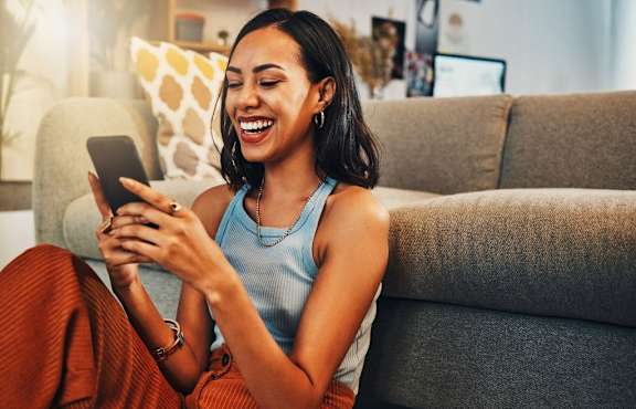 A woman is sitting on a couch and laughing while looking at her phone.