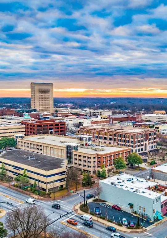 A cityscape with a mix of modern and older buildings, a clear sky, and a sunset in the distance.