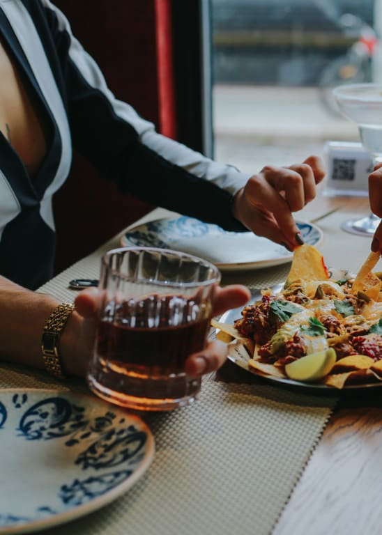 A person is serving food to another person at a table with a plate of nachos and drinks.