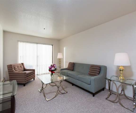 The spacious living room with a gray couch, brown throw pillows, wall art, brown plush chair, glass coffee table, decorations, a TV, lamps, and glass doors to the balcony at the Outlook Apartment Homes. 