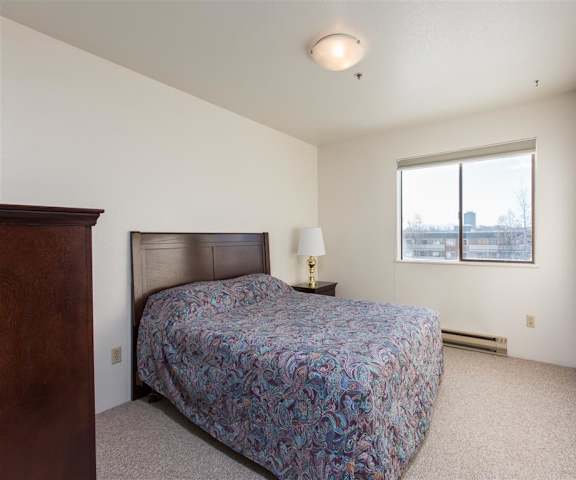 A spacious bedroom with natural lighting, a queen-size bed with a bedspread, a dresser, wall art, lamps, decorations, window, and nightstand with a lamp at the Outlook Apartment Homes. 