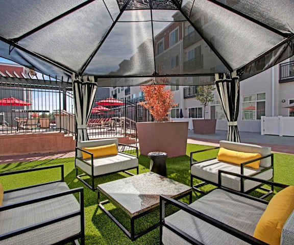The outdoor patio with a sheer black tent, white plush outdoor chairs, a coffee table, on the grassy with a fenced off pool and the apartment buildings in the background at the Ritz Classic Apartment Homes. 