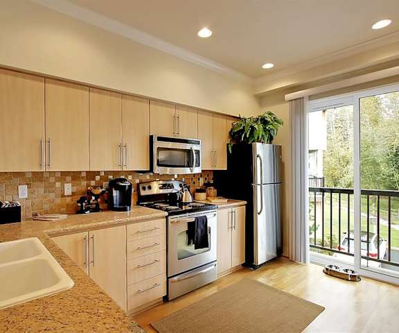 2-Bedroom Apartments in Kenmore, WA - Northshore Townhomes - Kitchen with Hardwood Flooring, Stainless Steel Appliances, Tan Wood-Tone Cabinetry, Slab Granite Countertops with Tile Backsplash, Decorative Plants, and Sliding Glass Door with Balcony Access.