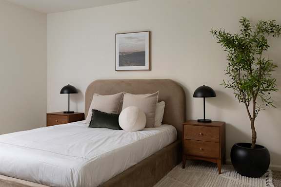 A bedroom with a large bed, two lamps, a potted plant, and a framed picture on the wall.