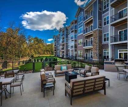 The outdoor patio with chairs and couches with beige cushions, a grill, glass firepits, coffee tables, and dining tables and chairs at Oaks of Shorewood Apartments. 
