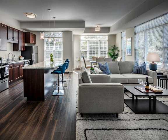 The living room with large windows, a couch, plush chairs, coffee table, decorations, a TV, and view of the dining room and the kitchen with beautiful dark wood cabinets, marble countertops, and stainless-steel appliances at Oaks of Shorewood Apartments. 