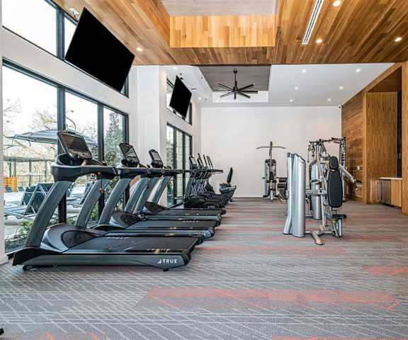 A gym with cardio machines, windows, TVs, and weights at Magnolia on the Green Apartments. 